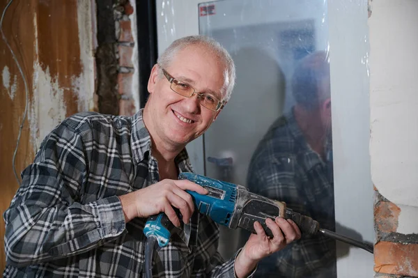 Portrait of a man 55-60 years old in a working environment during the repair of an apartment with his own hands, in a plaid shirt, smiling, wearing glasses, holding a perforator Royalty Free Stock Photos