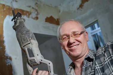 55-60 yaşlarında bir adamın portresi. Bir apartmanın tamiri sırasında kendi elleriyle, ekose gömlekli, gülümseyen, gözlük takan, bir delgi tutan bir adam.
