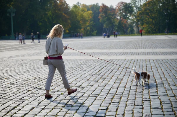 Young blonde woman in light casual clothes walks with a Cavalier King Charles Spaniel dog in the historical center of the small town of Kronstadt on a sunny fine day along the cobblestones — Stock Photo, Image