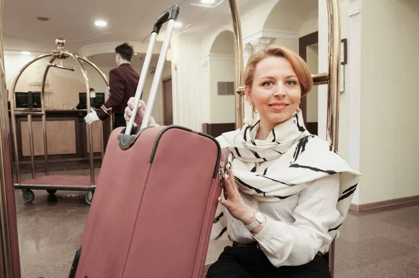 35-40-летняя женщина останавливается в отеле. Блондинка у тролля для перевозки чемоданов и багажа. В летней одежде. Она улыбается. Хорошее настроение — стоковое фото