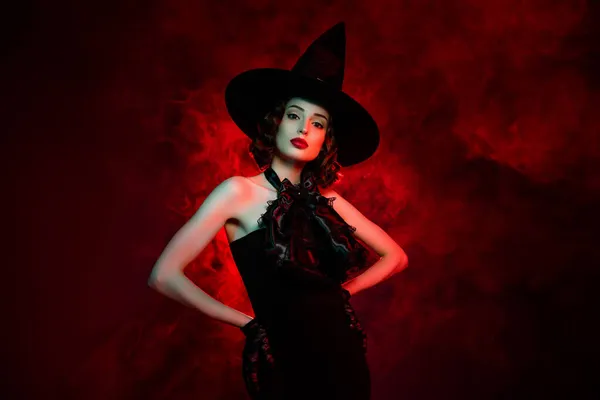 Foto de assustador adorável misterioso mulher desgaste gótico bruxa vestido headwear braços cintura isolado vermelho nevoeiro cor fundo — Fotografia de Stock