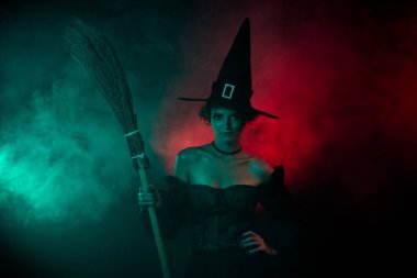 Büyüleyici kadın büyücünün elinde süpürge sopası karnaval hazırlığı. Canlı neon rengi arka planda izole edilmiş..