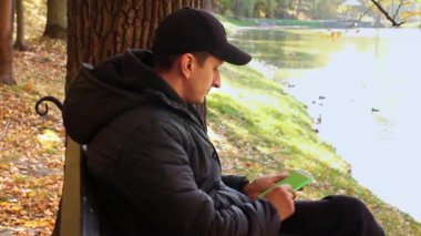 Genç bir adam sonbaharda parkta bankta oturuyor ve telefonda yeşil ekrana bakıyor, internette krom tuşuna basıyor sosyal medyayı okuyarak ellerini kapatıyor. Elleri göster.