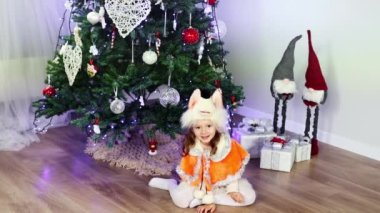 A small curly girl of European appearance in a fox costume sitting under the Christmas tree and looking at the camera and smiling in slow motion. New Years masquerade. Concept of Christmas and New