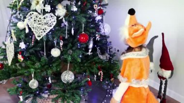 A small curly girl of European appearance in a fox costume dances and tells something near the Christmas tree in slow motion. New Year's masquerade. Concept of Christmas and New Year