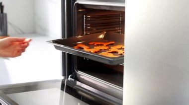 The womans hands open the oven and stick the baking sheet with ginger biscuits there. A young woman in a white modern kitchen cooking food in an oven close up. Concept of Christmas and New Year.