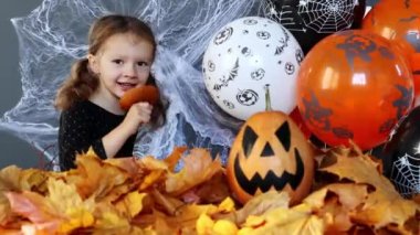 A little witch girl is funny eating a slice of pumpkin on the background of Halloween attributes. Halloweens holiday attributes. Lantern carved from pumpkin known as Jack-o-lantern. Trick or treat.