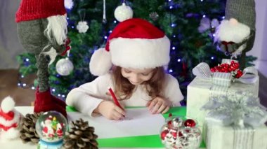 Kırmızı Noel Baba şapkalı küçük bir kız Noel Baba 'ya Noel ağacının altına dilekleriyle bir mektup yazıyor. Noel ve Yeni Yıl atmosferi.