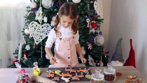 幸せな女の子はスローモーションで自宅でクリスマスジンジャーブレッドクッキーを着色します 台所でクリスマスの装飾 妖精の光を持つモミの木 新年とクリスマスの概念 — ストック動画