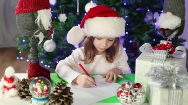 A little girl in a red Santa's Hat writes a letter to Santa Claus with her wishes under the Christmas tree. Christmas and New Year atmosphere.