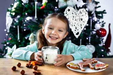 Kıvırcık saçlı küçük bir kız bardaktan süt içer ve bir Noel ağacının arka planında mutlu bir şekilde güler. Noel ve Yeni Yıl atmosferi.