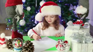 A little girl in a red Santa's Hat writes a letter to Santa Claus with her wishes under the Christmas tree. Christmas and New Year atmosphere.