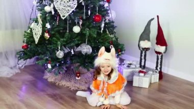 Tilki kostümü giymiş, Noel ağacının altında oturan ve kameraya bakıp gülümseyen Avrupalı kıvırcık bir kız. Yeni yıl maskeli balosu. Noel ve Yeni Yıl kavramı.