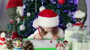 Kırmızı Noel Baba şapkalı küçük bir kız Noel Baba 'ya Noel ağacının altına dilekleriyle bir mektup yazıyor. Noel ve Yeni Yıl atmosferi.