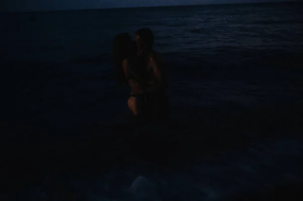 Nahý pár v lásce dívka a muž objetí a polibky ve vodě v moři na pláži v letní noci — Stock fotografie