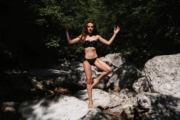 Young sexy brunette girl in a black swimsuit stands in yoga pose on rocks by river in nature — Stock Photo, Image