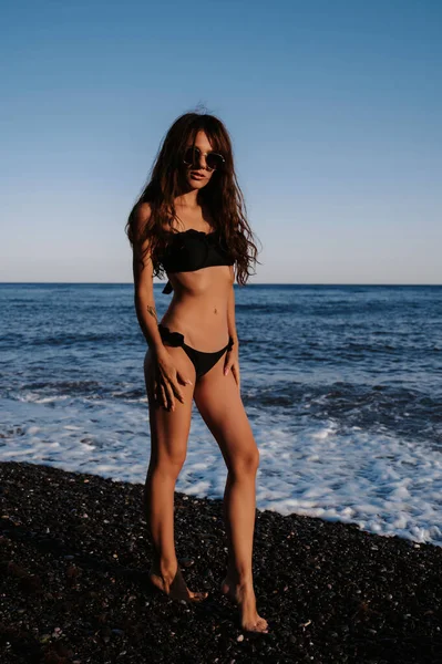 Sexy chica morena en un traje de baño negro con una figura delgada posando en la playa junto al mar — Foto de Stock