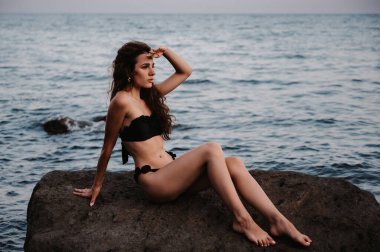 Yazın deniz kenarındaki kayalıklarda oturan siyah mayolu seksi genç kız.