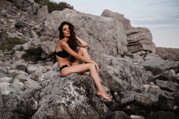 Sexy young girl in black swimsuit sitting on the rocks by the sea in summer — Stock Photo, Image
