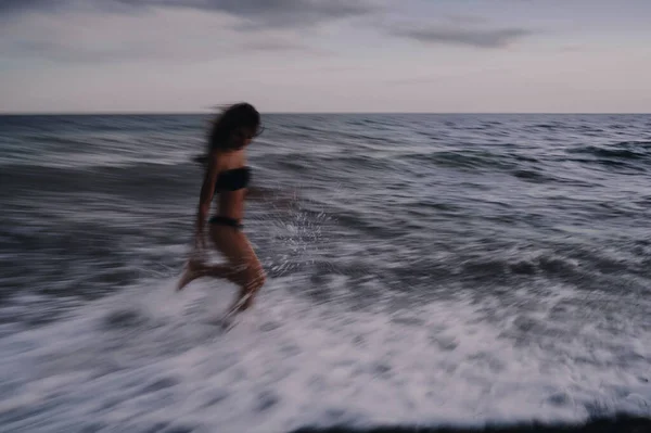 Šťastná dívka v plavkách šťastně běží podél moře vlny na pláži v letním večeru — Stock fotografie