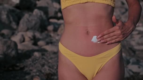 Woman with tanned skin in a swimsuit applies sunscreen to her tummy in summer on the beach — Wideo stockowe