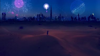 AERİAL. Geleneksel Arap elbisesi içinde genç bir kadın - abaya çölde yükselişte duruyor ve modern Dubai şehrindeki fantastik havai fişek gösterisine bakıyor..