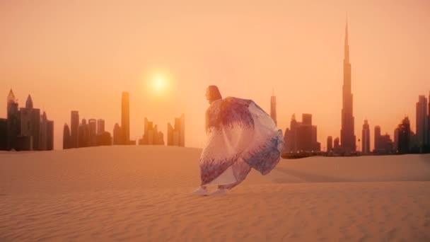 Mulher árabe vestida em vestido tradicional dos Emirados Árabes Unidos abayain levantando as mãos no pôr do sol em um deserto com silhueta da cidade de Dubai no fundo. — Vídeo de Stock