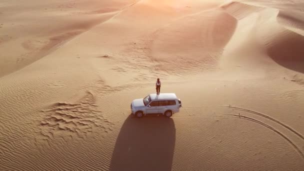 Letecký pohled na ženskou cestovatelku stojí na střeše auta a zvedá ruce na západ slunce v poušti. — Stock video