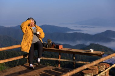 Asyalı yaşlı kadın portresi, ahşap bankta oturuyor, kahve setiyle ve el kaldırarak, dağ veya tepenin görüş açısını kapatıyor, mutlu emeklilik kavramı, kopyalama alanı.