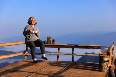 Asyalı yaşlı bir kadının portresi, tahta bankta oturuyor. Elinde kahve, dağ veya tepenin manzarası, mutlu emeklilik konsepti, kopyalama alanı.