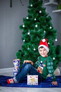 Geyik ve Noel Baba şapkalı yeşil kazaklı bir çocuk Noel ağacının arka planında hediyelerle oturuyor.