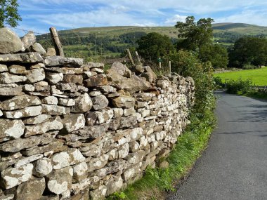 Westholme Yolu 'nun köşesinden aşağı doğru uzanan büyük kuru taş duvar. Uzak köşelerinde tarlalar, ağaçlar ve tepeler var. Leyburn, Yorkshire, İngiltere.
