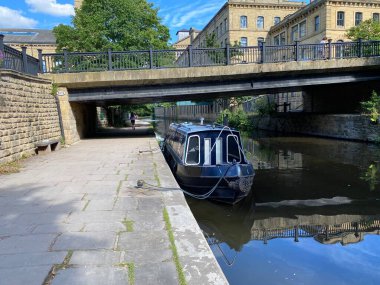 A view of the Leeds to Liverpool canal, with a boat, and old mill buildings in the background in, Saltaire, Bradford, UK
