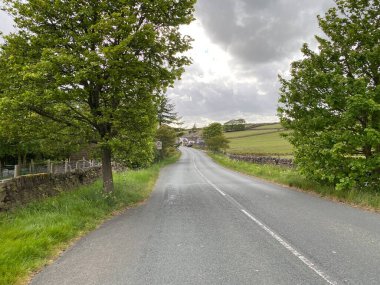 Long Causeway road, with trees, dry stone walls, and houses in the distance in, Denholme, Bradford, Yorkshire, UK