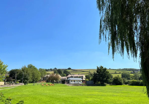 Looking toward a cricket pavillion, with trees and houses nearby, in the picturesque village of, Bradley, Skipton, UK