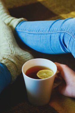 Limonlu bir fincan çay, bir kızın ellerinde. Mavi kot pantolon ve örgü çorap giymiş bacakların yakın çekimi. Yakın plan, dikey fotoğraf.