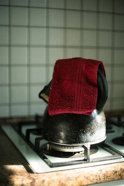 Bir yaz sabahı hafif bir ev mutfağının içinde. Siyah bir çaydanlık elinde kırmızı havluyla gaz sobasının üzerinde duruyor. Güneş ışığı düşüyor.