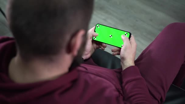 Hombre en la habitación moderna sentado en un sofá usando el teléfono con pantalla verde. Hombre relajado mirando el teléfono inteligente con pantalla verde simulada croma pantalla clave — Vídeo de stock