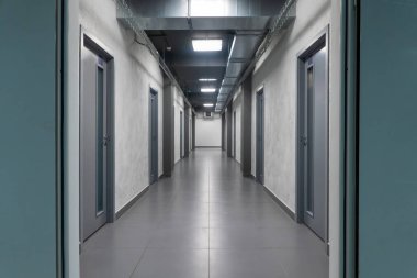 Birkaç gri kapılı, uzun, modern, boş ofis koridoru manzarası. Zemin kiremitli ve ışıklar açık.. 