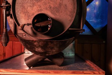 Close-up of a round heating stove near the window. There is rust and scale on the steel surface. A fire is visible in the blower. Background. 