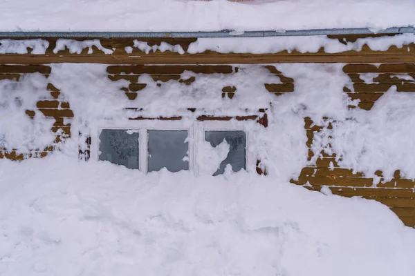 A fragment of the wall of a wooden house covered with snow in the middle of a small window. The glass is covered with frost. Details of winter village life. Background. 
