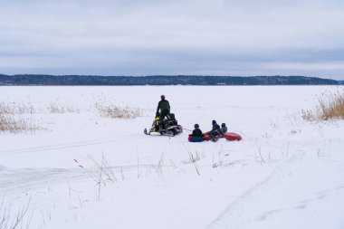Winter landscape of a large lake covered with ice and snow. You can see the figure of a man on a snowmobile, which rolls several children on tubing. 