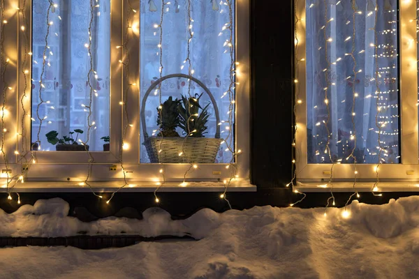 Ahşap bir evin penceresi şenlikli sarı bir çelenkle süslenmiştir. Kar pencere eşiğine ulaşır. İçeride kapalı bitkiler var. Yakın plan. Arkaplan. 