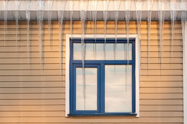 Pencereli bej kaplamalı bir ev duvarı parçası. Çatıda büyük buz sarkıtları asılı. Camda gölgeler ve yansımalar var. Arkaplan 