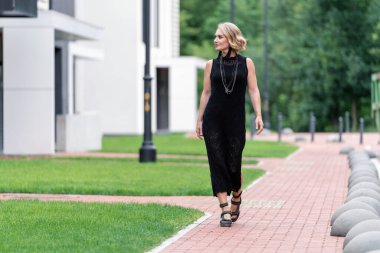 Beyaz, beyaz, yetişkin, siyah elbiseli ve boynunda bijouterie olan güzel bir kadın. Güzel Yeşil Çimen ve Binalar Arkaplanda Mimari.