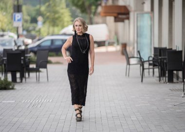 Litvanya 'daki Vilnius Old Town Caddesi. Beyaz, beyaz, yetişkin bir kadın siyah elbise ve boynuna bijouterie giyiyor. Kaldırımda yürüyorum. Dışarıdaki Sandalyelerle Arka planda Dükkan ve Restoran