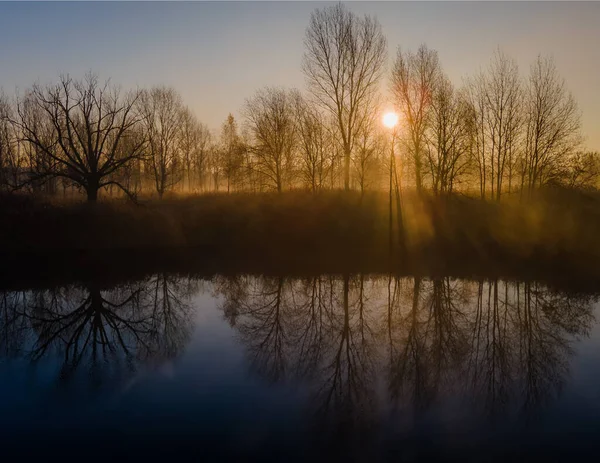 Promienie porannego słońca świecące wśród jesiennych drzew i odbite w wodzie mglistego jeziora. — Zdjęcie stockowe