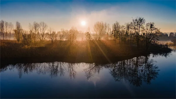 Les rayons du soleil du matin brillent à travers les arbres d'automne et se reflètent dans l'eau du lac brumeux. — Photo