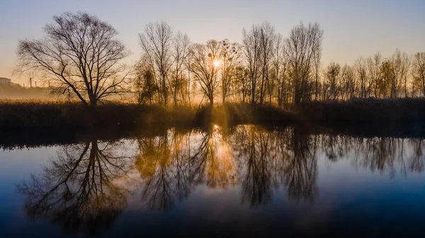 Les rayons du soleil du matin brillent à travers les arbres d'automne et se reflètent dans l'eau du lac brumeux. — Photo