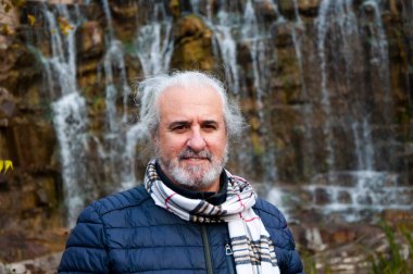 Sonbahar ceketi ve şelalenin arka planında eşarbı olan kır saçlı yaşlı bir adam..